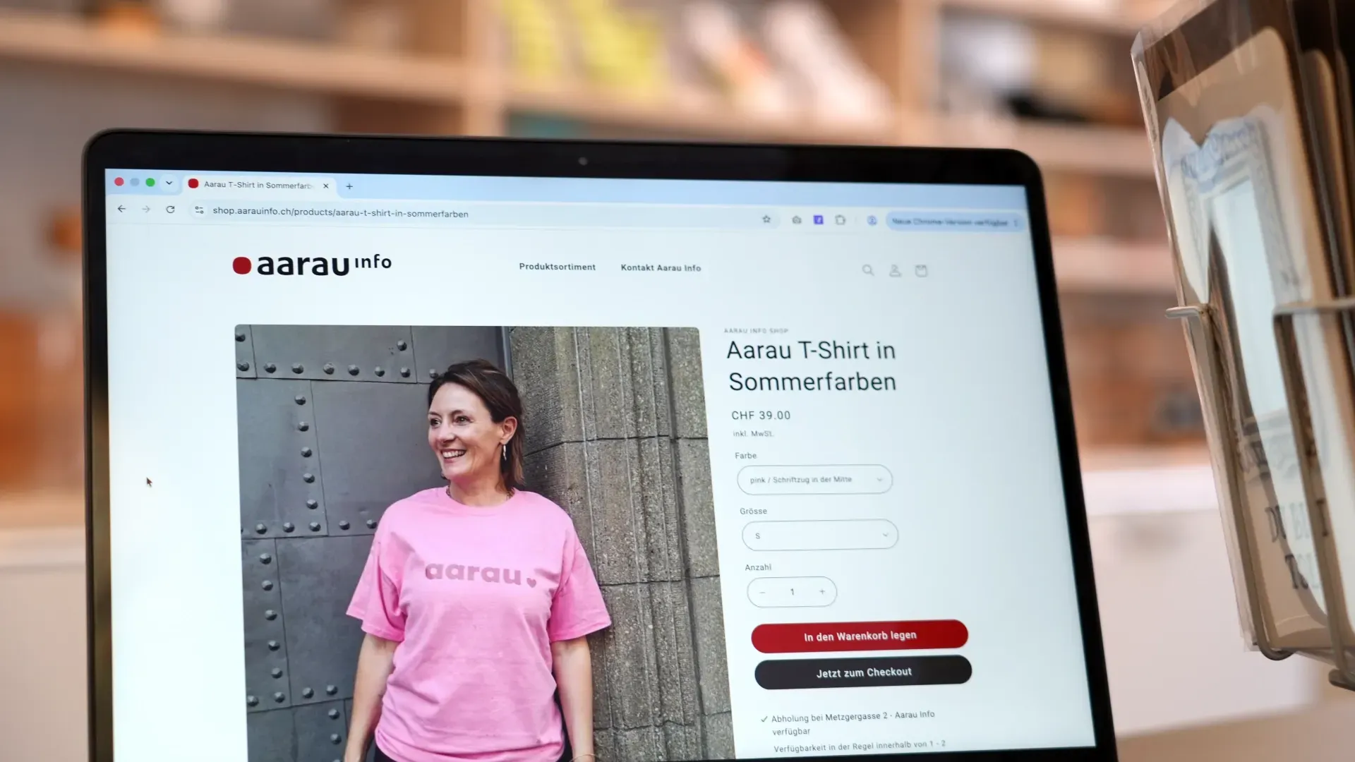 Laptop mit geöffneter aar au-info Produktseite; ein Modell im rosa T‑Shirt steht vor einer grauen Wand, daneben steht „Aarau T‑Shirt in Sommerfarben“. Rechts ein Smartphone im Halter.