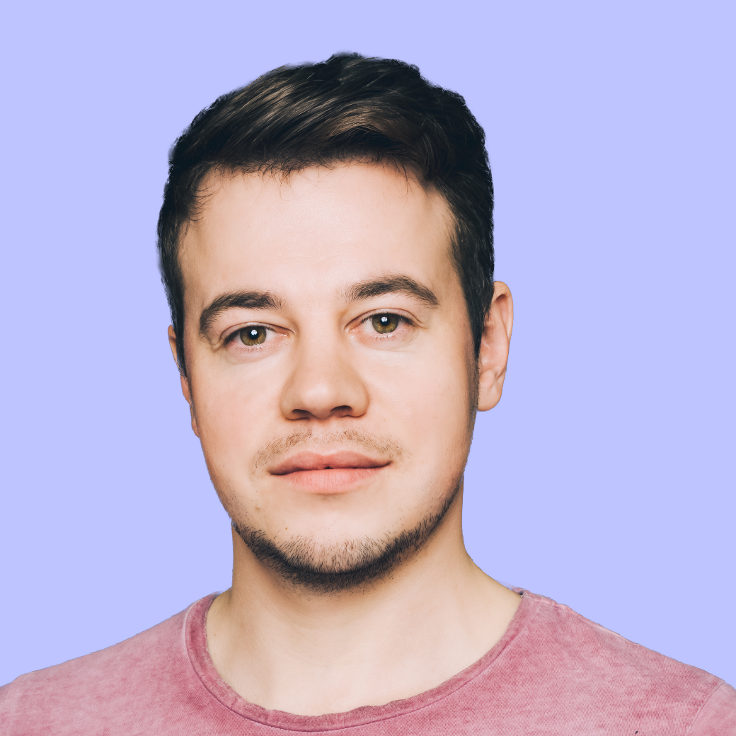 Porträt eines Mannes mit kurzen, dunklen Haaren und einem leichten Bart, der in die Kamera schaut. Er trägt ein rosa T-Shirt und hat einen neutralen Gesichtsausdruck. Der Hintergrund ist einfarbig in hellem Lila.