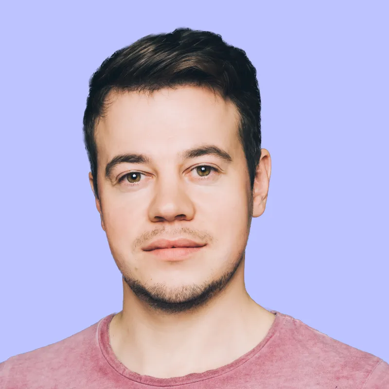 Porträt eines Mannes mit kurzen, dunklen Haaren und einem leichten Bart, der in die Kamera schaut. Er trägt ein rosa T-Shirt und hat einen neutralen Gesichtsausdruck. Der Hintergrund ist einfarbig in hellem Lila.