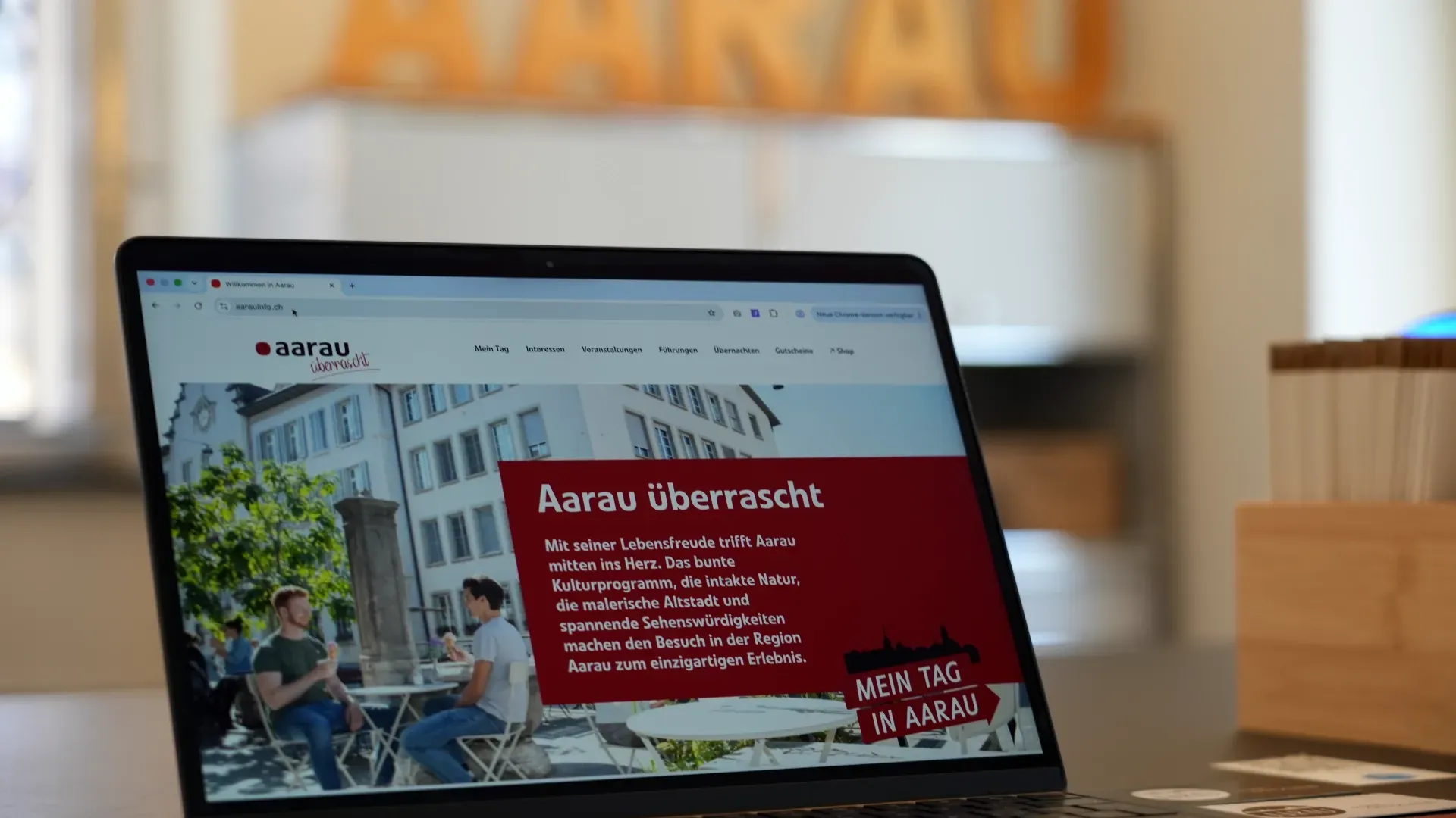 Laptop mit geöffneter Website „aarau“: rotes Banner „Aarau überrascht“, Text und Bild mit Personen auf einer Terrasse in einem hellen Raum.