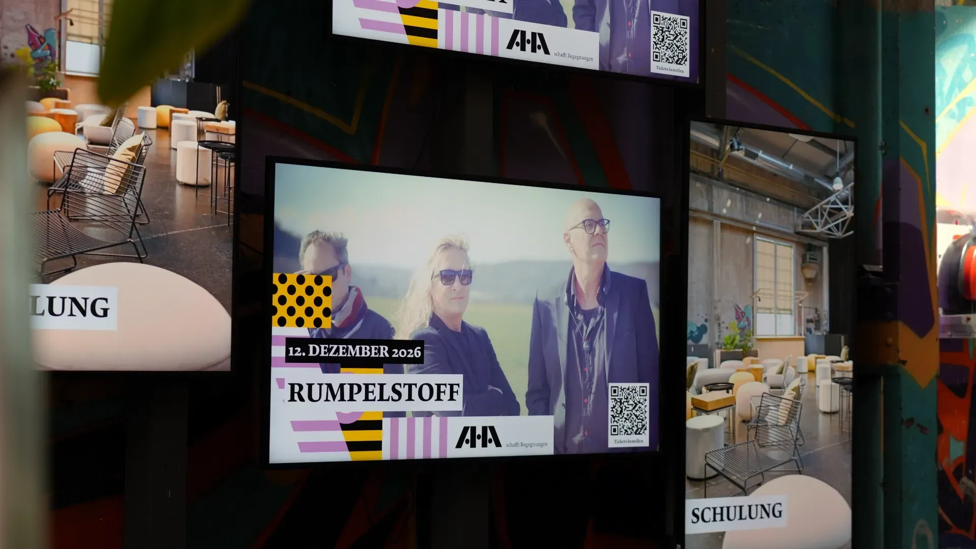 Mehrere LED-Bildschirme in einem Raum, zentral ein Videoframe mit drei Personen, Text „12. DEZEMBER 2026“ und „RUMPelSTOFF“, daneben „SCHULUNG“ und „LUNG“.