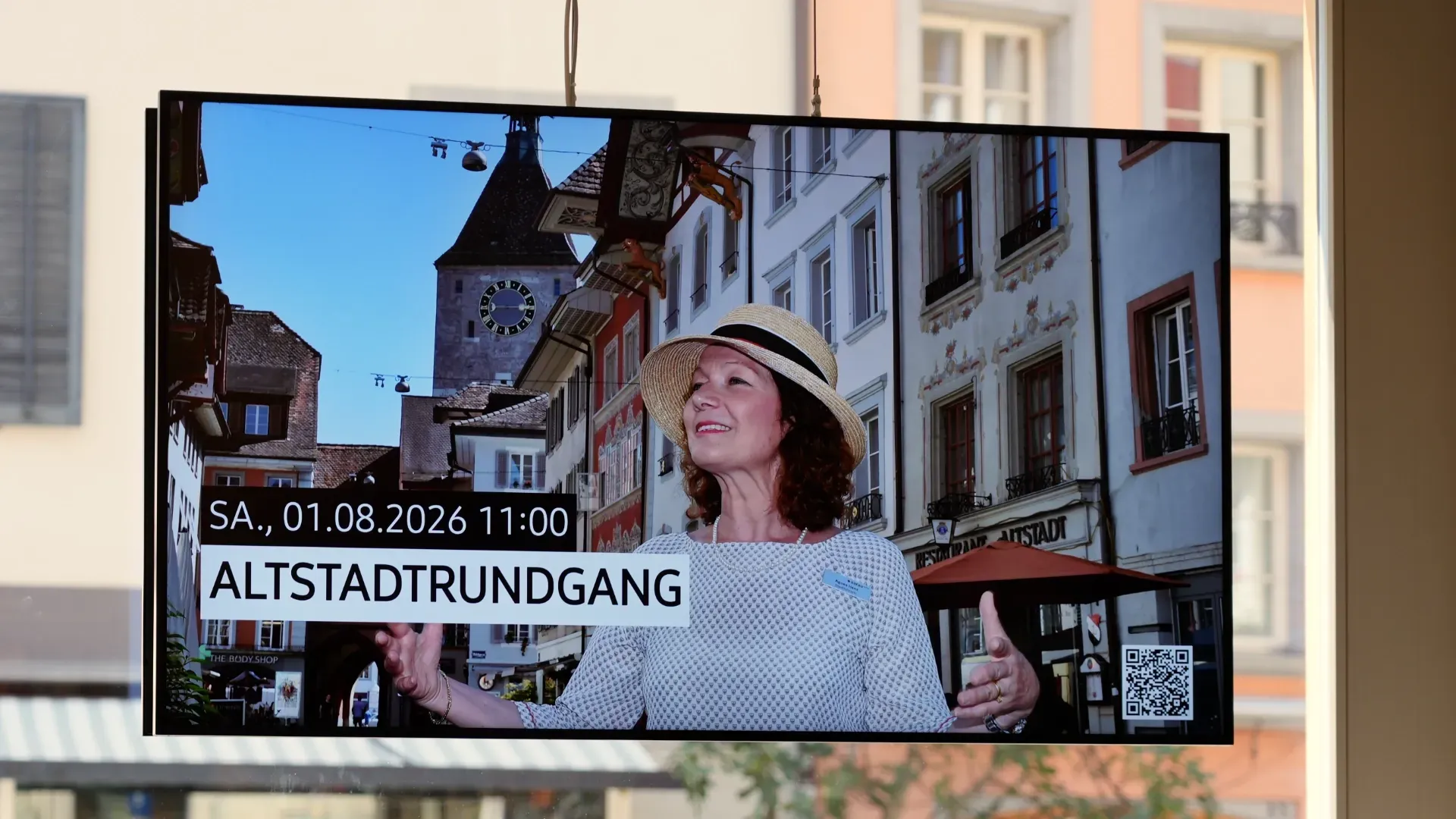 Frau mit Strohhut auf Stadtbildschirm: Altstadt-Rundgang, Datum und Uhrzeit eingeblendet; im Hintergrund Fachwerkhaus und Kirche, Häuserfassaden in der Altstadt.