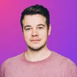 Porträt eines Mannes mit dunklem Haar und rosa T-Shirt vor pink-violettem Hintergrund.