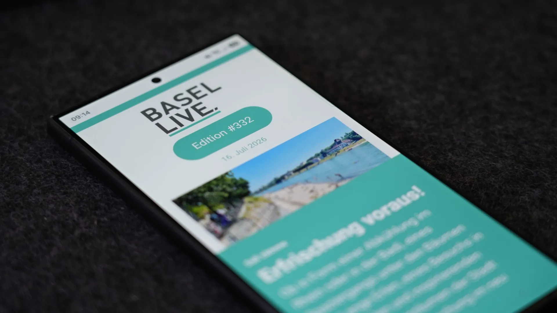 Smartphone mit Anzeige „Basel Live“, Edition #332, auf schwarzem Hintergrund, schräg liegend; grüner Header und Landschaftsfoto im oberen Bereich.