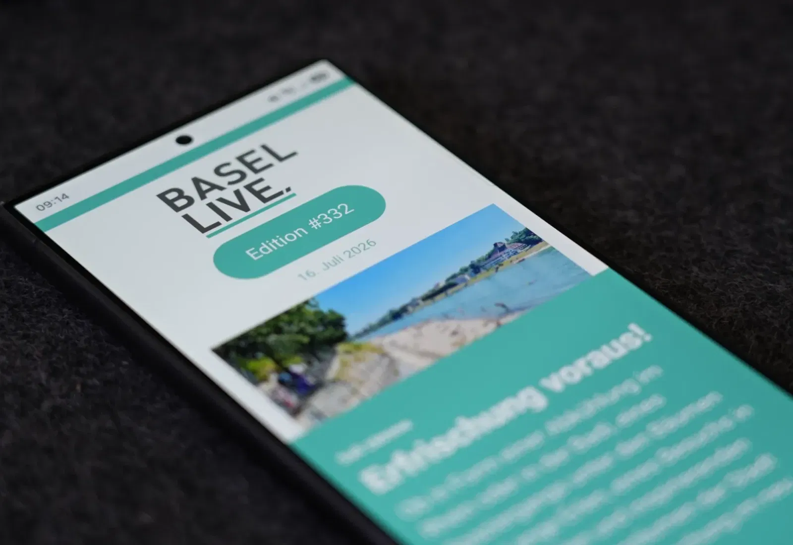 Smartphone mit Anzeige „Basel Live“, Edition #332, auf schwarzem Hintergrund, schräg liegend; grüner Header und Landschaftsfoto im oberen Bereich.