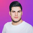 Porträt eines jungen Mannes mit dunklem Haar und Bart in weißem T-Shirt vor pink-violettem Hintergrund.