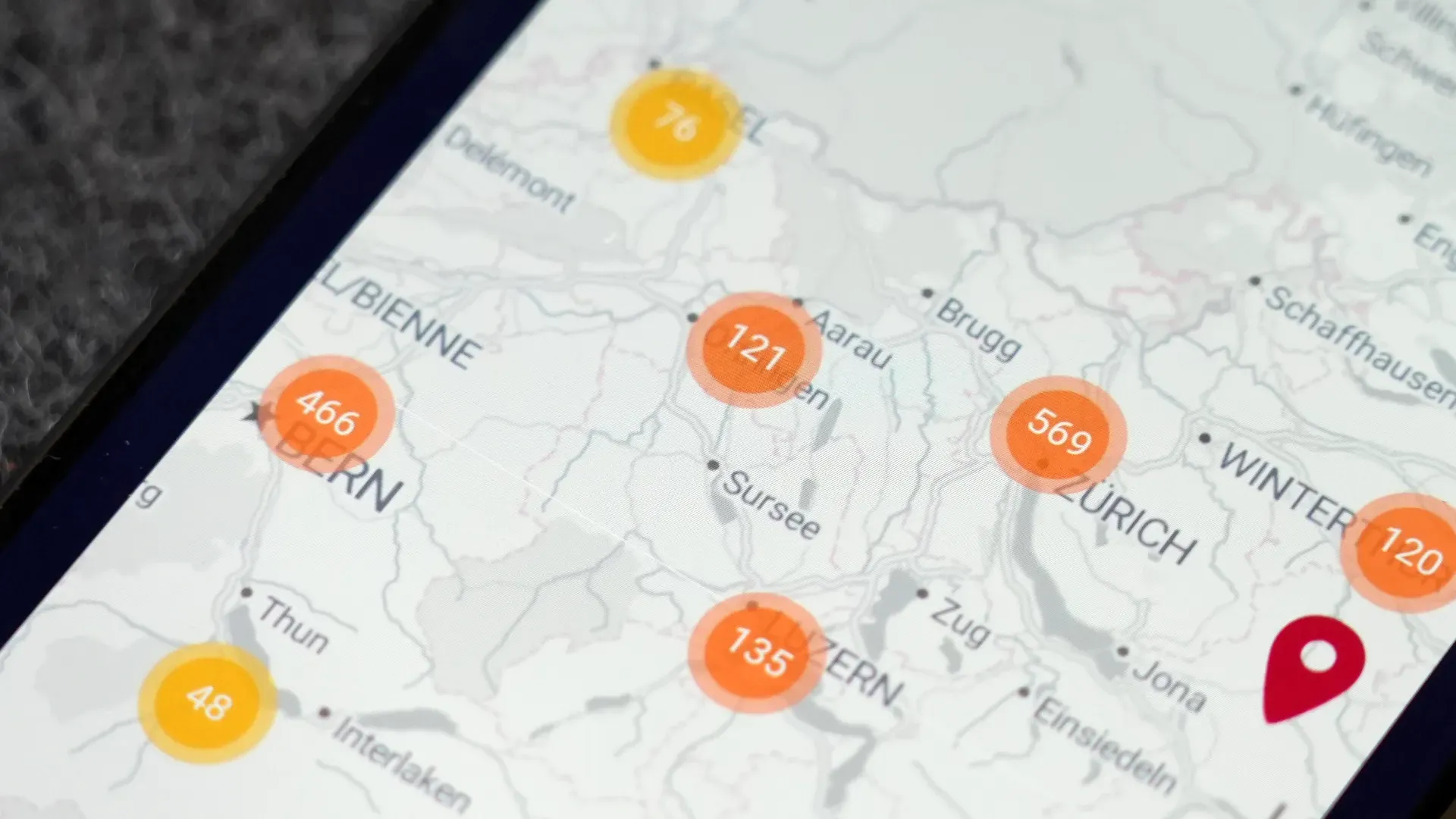 Landkarte der Schweiz mit orangefarbenen Markierungen und Zahlen sowie roter Standort-Pin, eingeblendet auf Smartphone-Display.
