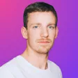Porträt eines Mannes mit kurzen dunklen Haaren und weißem T-Shirt vor pink-violettem Farbverlauf.