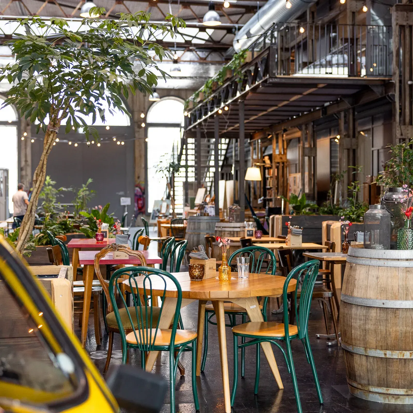 Innenhof einer Event- bzw. Gastronomiehalle mit Holztischen und Stühlen, Stehtischen in Holzbarrikaden, warmen Lichterketten, Pflanzen und einem Gelb-Schwarz-Taxi im Vordergrund.