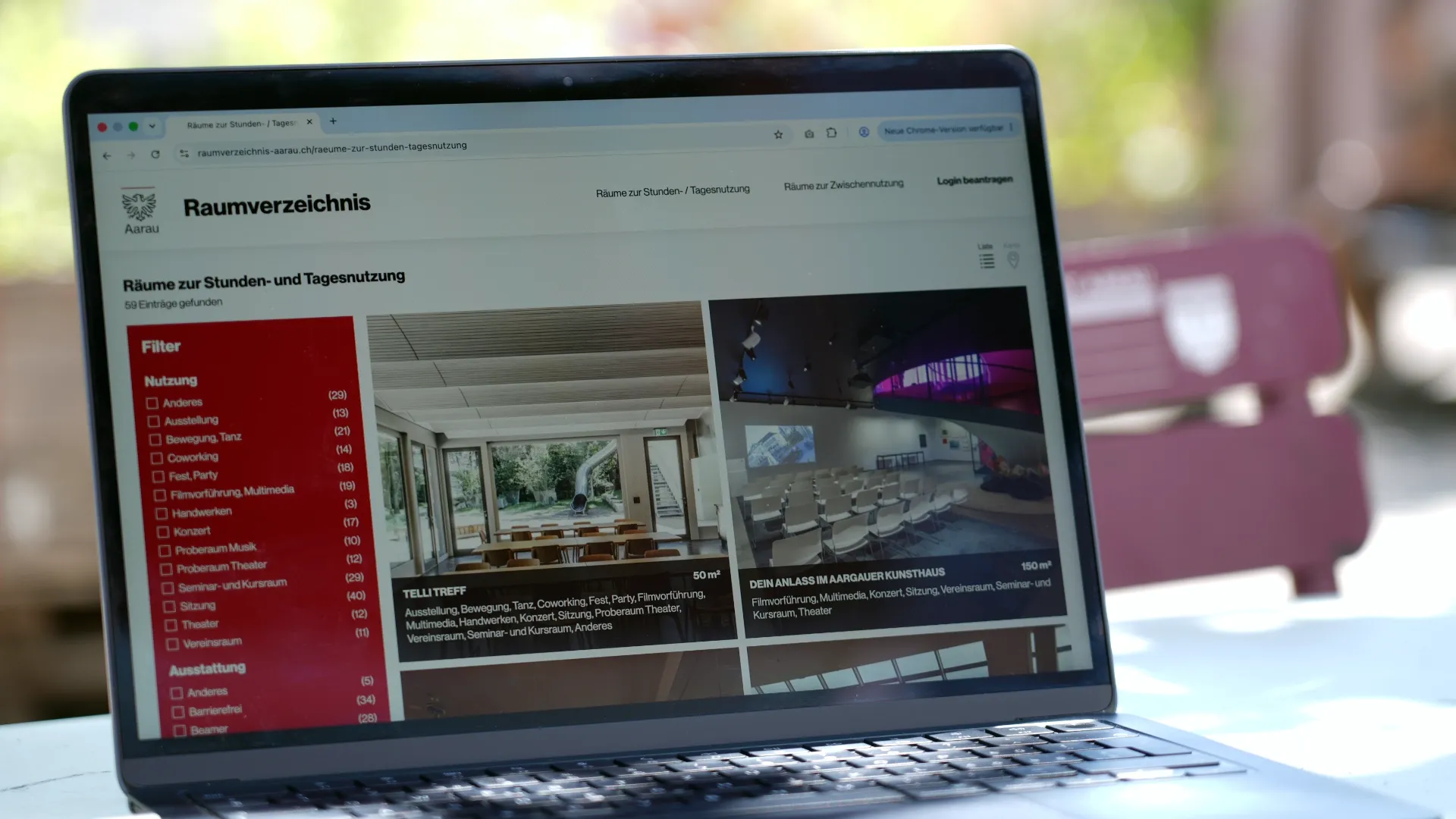 Laptop mit offenem Browser auf „Raumverzeichnis“, Liste links und Raster mit Raumansichten in Kacheln, unscharfer Hintergrund im Freien.