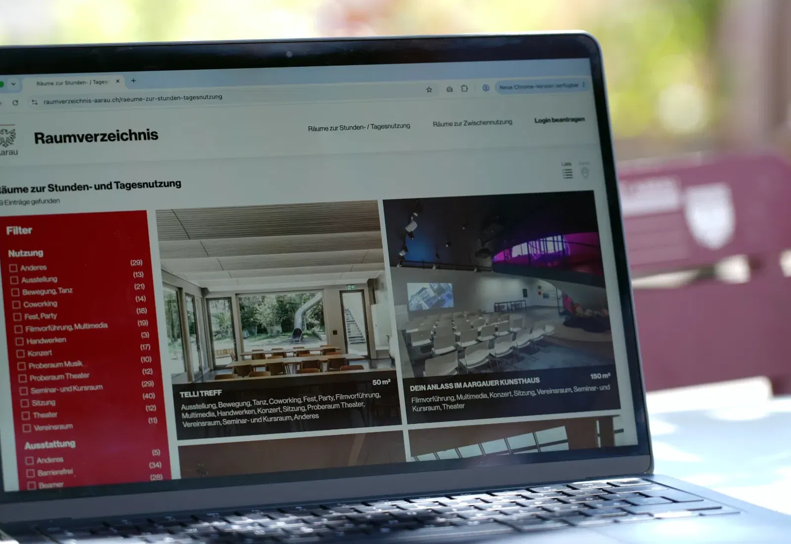 Laptop mit offenem Browser auf „Raumverzeichnis“, Liste links und Raster mit Raumansichten in Kacheln, unscharfer Hintergrund im Freien.