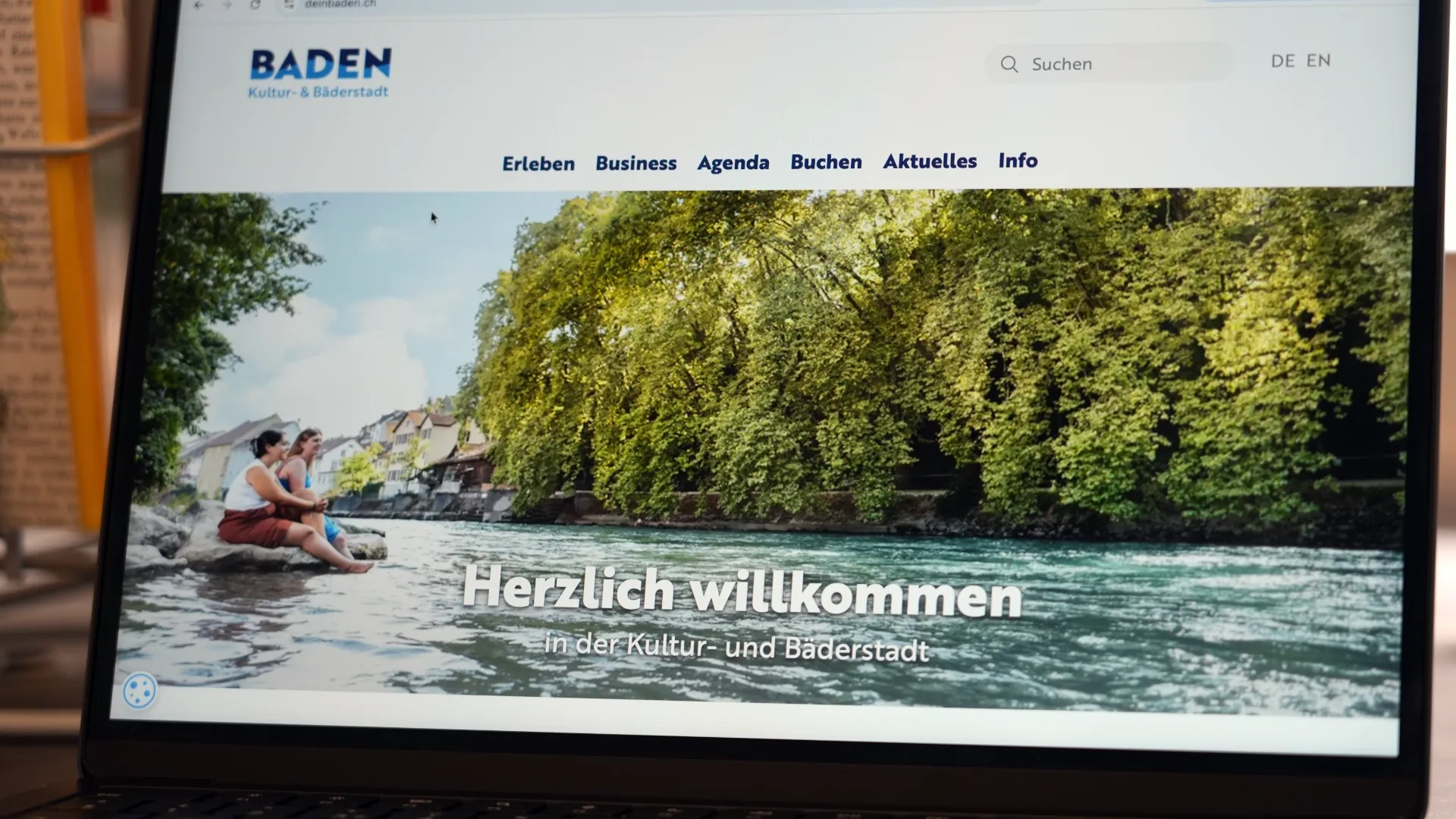Website „Baden“ auf einem Bildschirm: Flusslandschaft mit Ruderboot, Menschen im Boot, grünes Ufer und Überschrift „Herzlich willkommen“ mit Navigation oben.