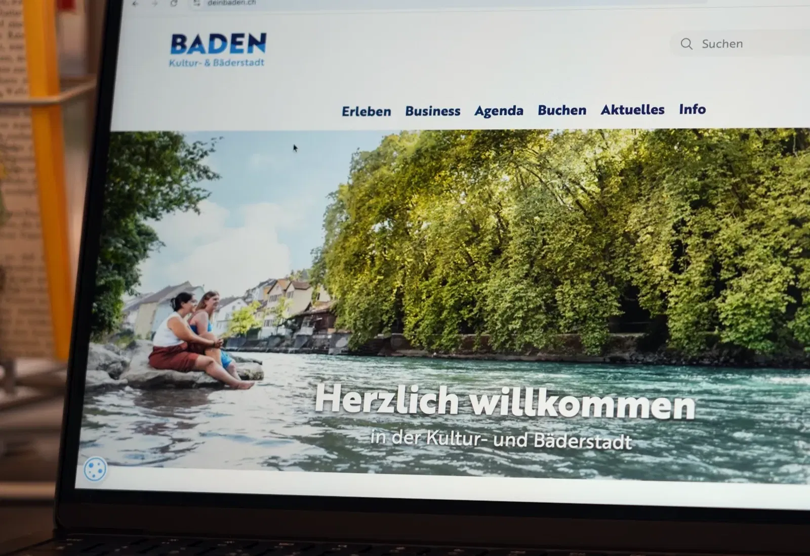 Website „Baden“ auf einem Bildschirm: Flusslandschaft mit Ruderboot, Menschen im Boot, grünes Ufer und Überschrift „Herzlich willkommen“ mit Navigation oben.