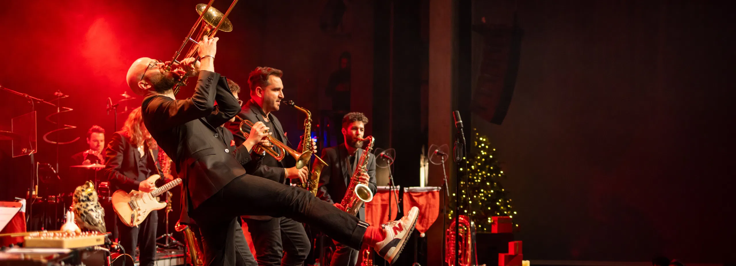Liveband auf beleuchteter Bühne: Sänger mit Trompete im Vordergrund, Musiker mit Gitarren und Saxofonen dahinter, Weihnachtsbaum und Publikum rechts.