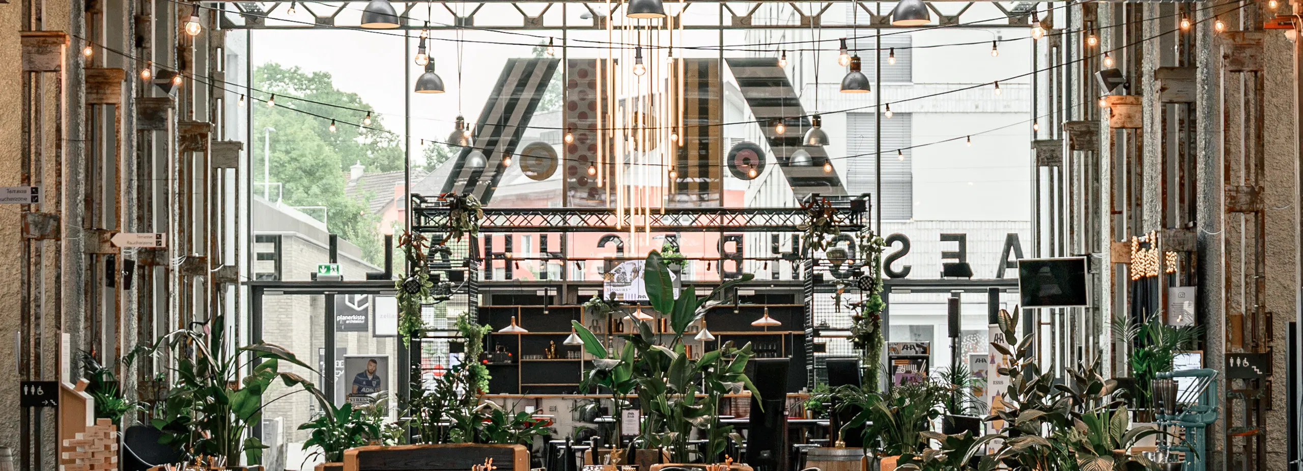 Industrielles Restaurant mit zahlreichen Holztischen, Stühlen, Hänge- und Lichterketten, sichtbaren Lüftungsrohren, großen Fenstern und üppigen Zimmerpflanzen.