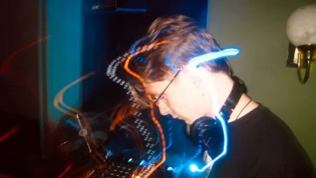 Person mit Kopfhörern und leuchtendem Stirnband in abgedunkelter Umgebung, vor farbig beleuchtetem Mischpult.