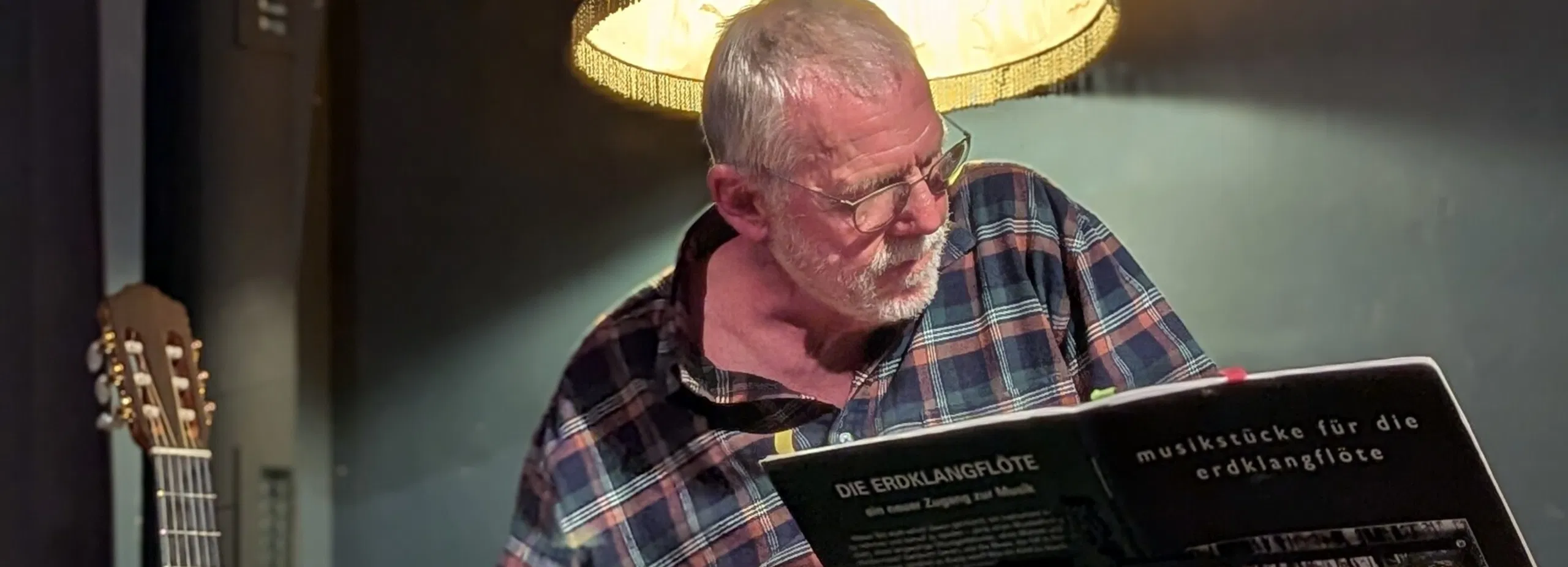 Älterer Mann spielt ein Saiteninstrument unter einer großen Lampe; daneben steht eine Gitarre, davor liegt ein Notenständer.
