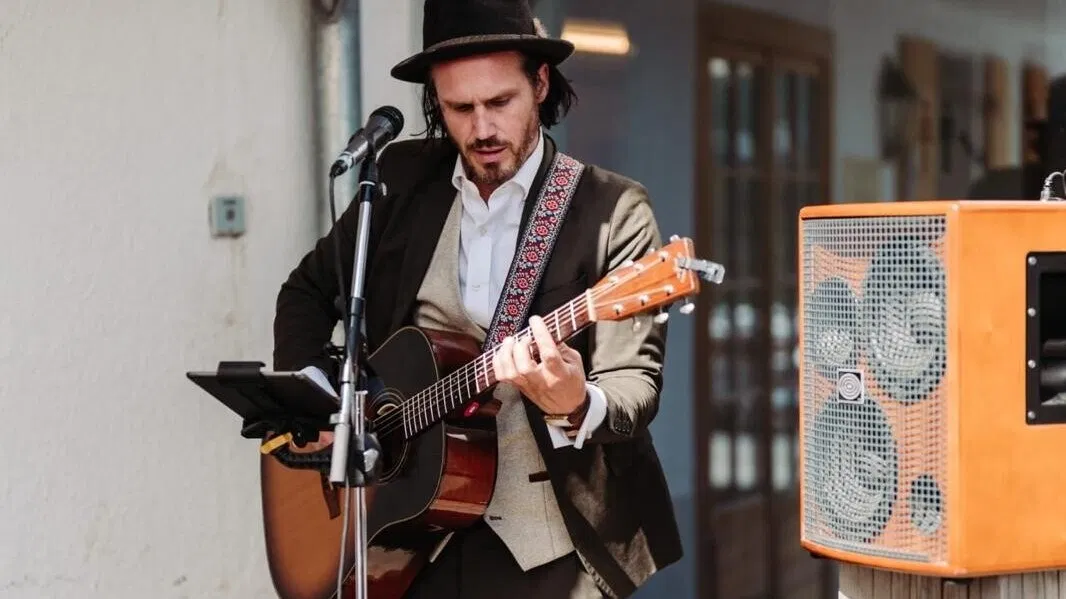Musiker mit Hut und Anzug spielt Akustikgitarre am Mikrofon; daneben stehen orangefarbener Lautsprecher und Verstärker auf einer Holzterrasse.