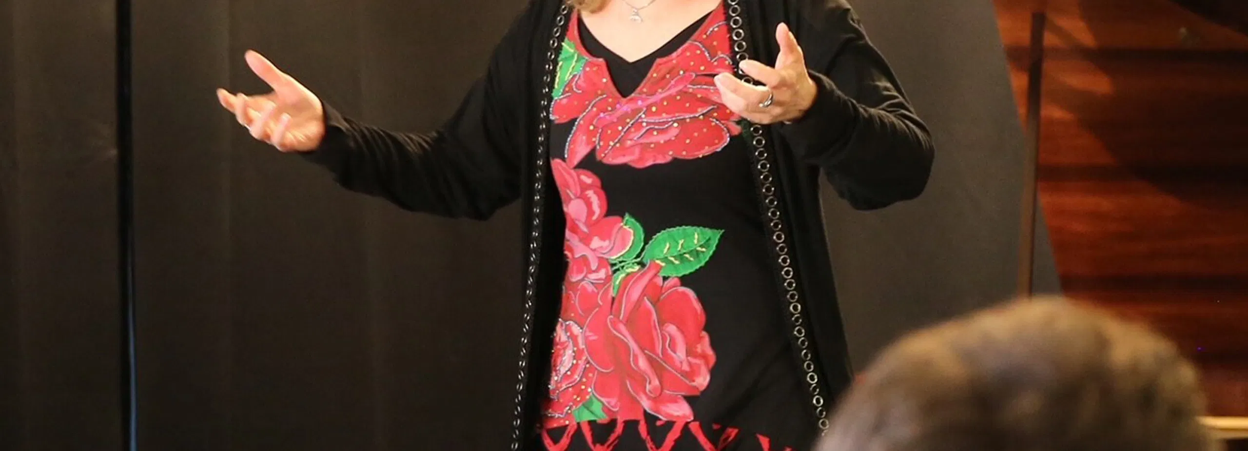 Frau in floralem Kleid spricht mit ausgestreckten Händen vor Publikum; im Hintergrund steht ein Klavier.