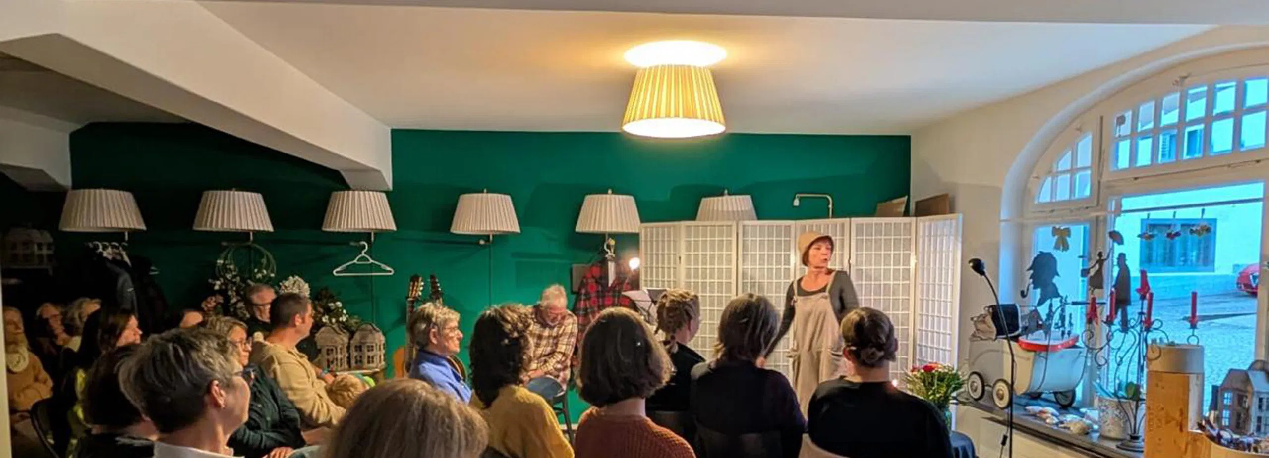 Publikum sitzt in einem gemütlichen Raum vor einer sprechenden Frau; grüne Wand, mehrere Lampen, großes Rundbogenfenster und dekoriertes Fensterbrett.