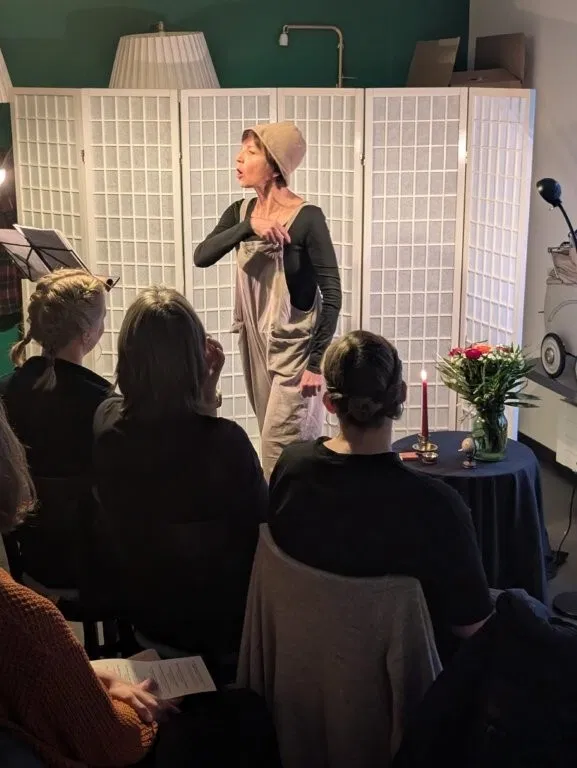 Schauspielerin in Kostüm spricht vor Publikum in einem Raum mit Paravent, Stehlampe, Mikrofon und gedecktem Tisch mit Blumen und Kerze.
