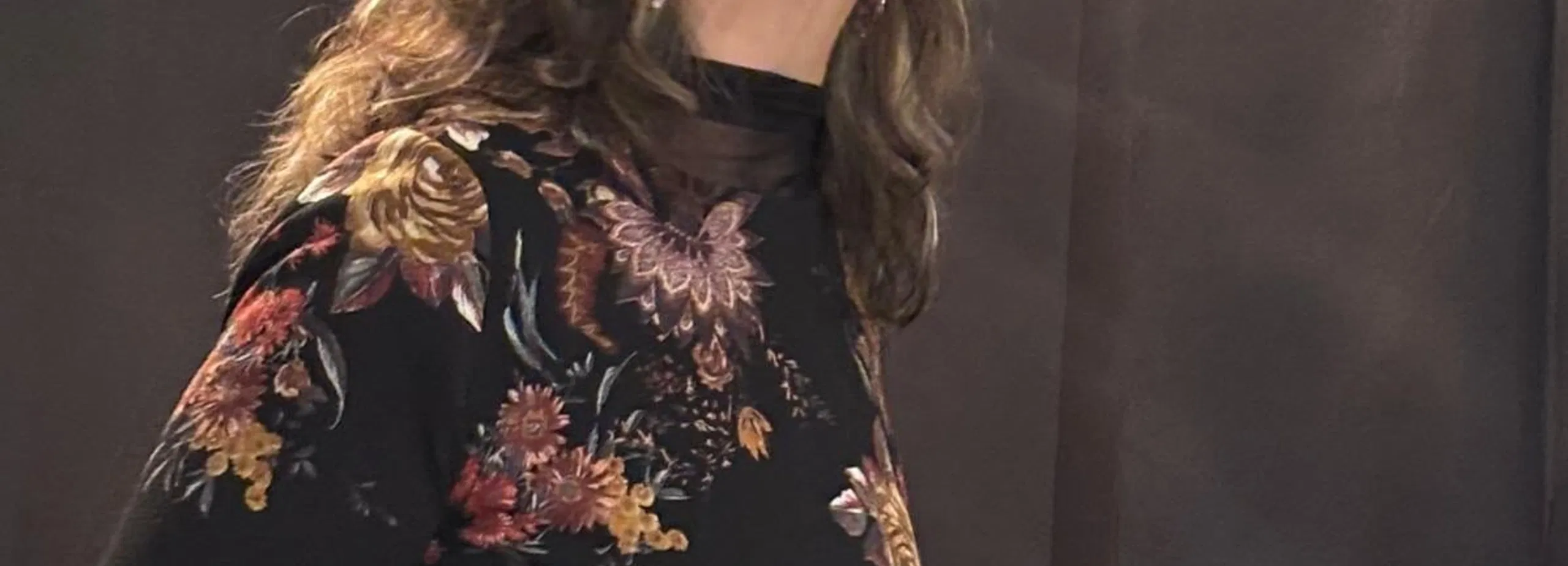 Frau mit langen braunen Haaren und floral gemustertem Kleid vor schwarzem Vorhang.
