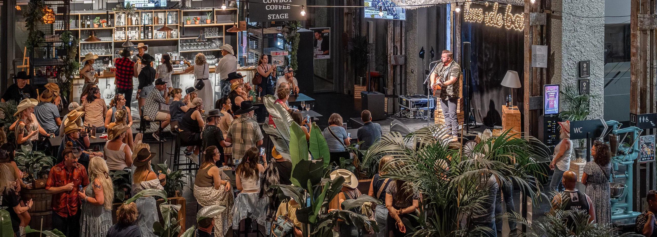 Innenraum eines belebten Lokals mit Cowboy-Coffee-Bühne, Musiker am Mikrofon, dichtem Publikum, Pflanzen, Bar und stimmungsvoller Beleuchtung.