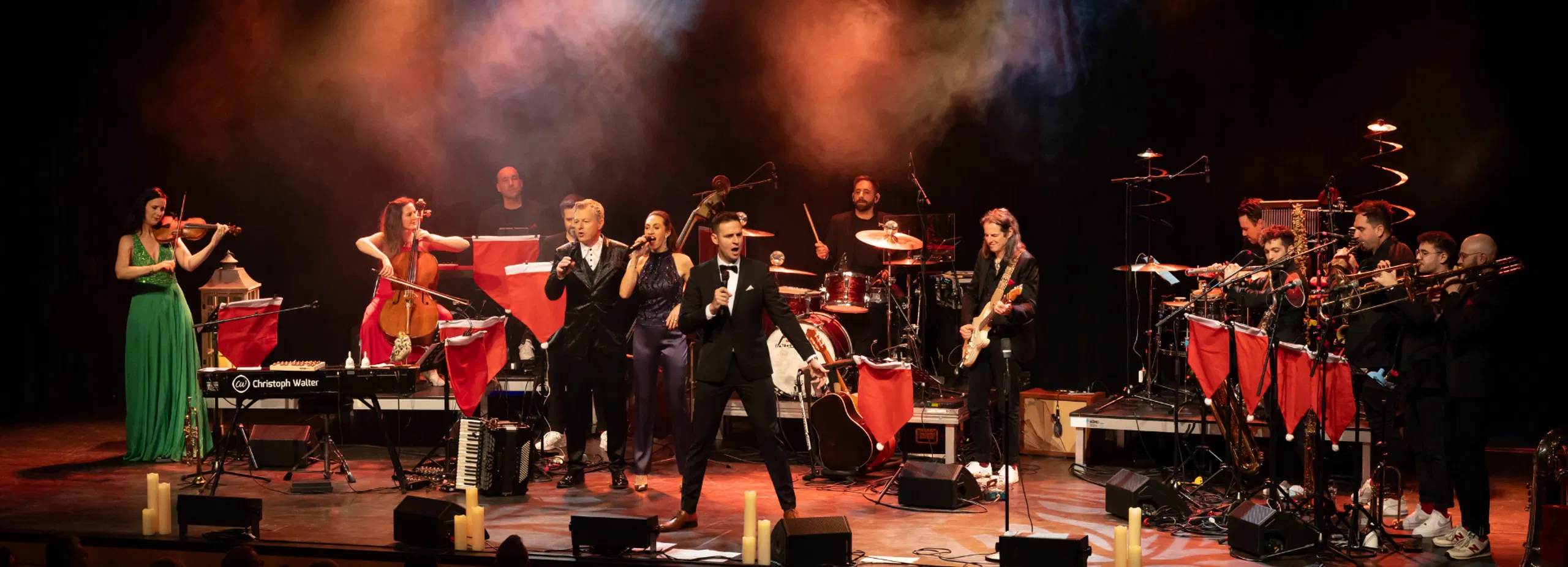 Großes Bühnenensemble mit Sängerinnen, Sängern und Instrumentalisten bei einem Konzert; farbige Scheinwerfer und Nebel, Publikum im Vordergrund.