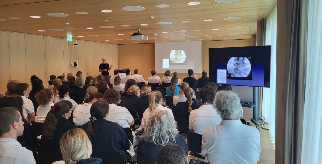 Ein Referent steht vor einer Gruppe von Zuhörern in einem modernen Seminarraum. Auf einer Leinwand sind medizinische Bilder zu sehen. Die Teilnehmer, überwiegend in weißen Oberteilen, sitzen in Reihen und richten ihre Aufmerksamkeit auf den Vortragende...