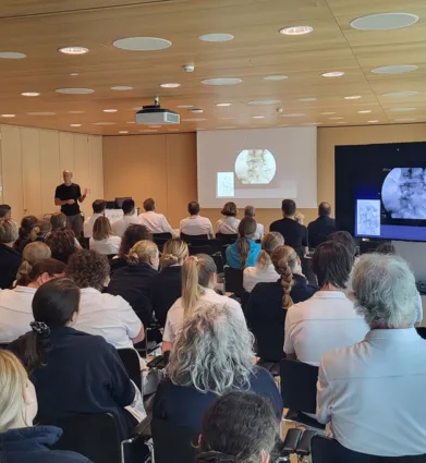 Ein Referent steht vor einer Gruppe von Zuhörern in einem modernen Seminarraum. Auf einer Leinwand sind medizinische Bilder zu sehen. Die Teilnehmer, überwiegend in weißen Oberteilen, sitzen in Reihen und richten ihre Aufmerksamkeit auf den Vortragende...