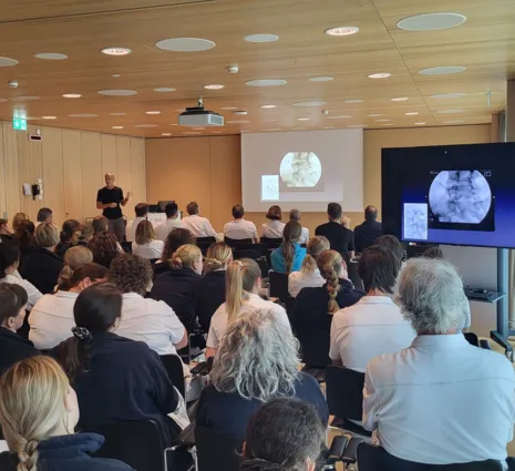 Ein Referent steht vor einer Gruppe von Zuhörern in einem modernen Seminarraum. Auf einer Leinwand sind medizinische Bilder zu sehen. Die Teilnehmer, überwiegend in weißen Oberteilen, sitzen in Reihen und richten ihre Aufmerksamkeit auf den Vortragende...