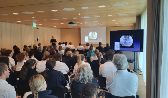 Ein Referent steht vor einer Gruppe von Zuhörern in einem modernen Seminarraum. Auf einer Leinwand sind medizinische Bilder zu sehen. Die Teilnehmer, überwiegend in weißen Oberteilen, sitzen in Reihen und richten ihre Aufmerksamkeit auf den Vortragende...