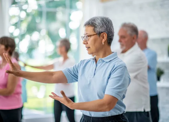 A group of seniors spend an afternoon indoors in a health center. They are doing group tai chi together.