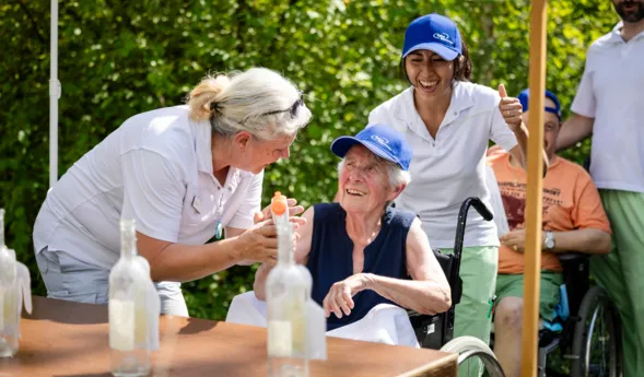 Betreuerinnen in blauen Kappen servieren Getränk und Wasserflaschen an eine ältere Frau im Rollstuhl bei einer Outdoor-Feier im Grünen.