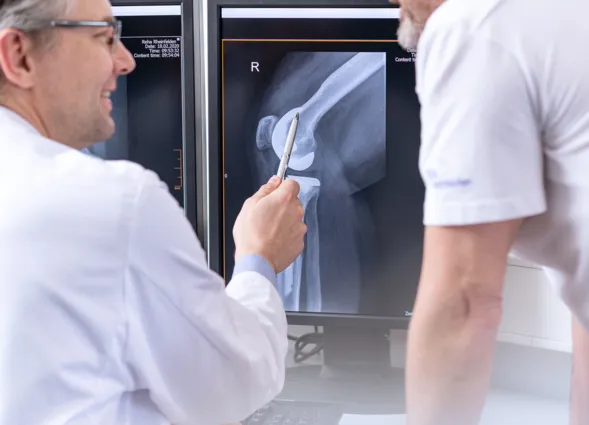 Zwei Ärzte in weißen Kitteln betrachten ein Röntgenbild eines Knies auf Computerbildschirmen. Einer der Ärzte zeigt mit einem Stift auf das Bild, während der andere aufmerksam zuhört. Ein Computer und eine Tastatur sind im Vordergrund sichtbar.