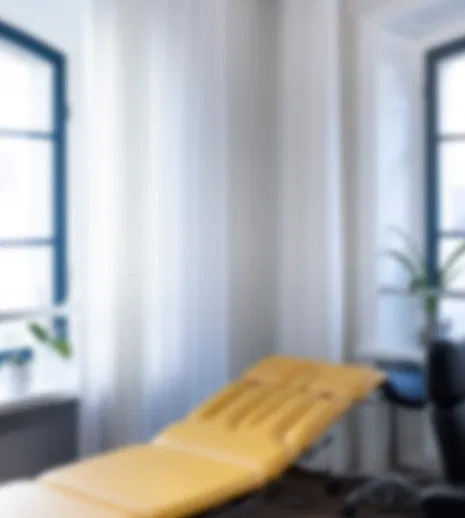 Heller Behandlungsraum mit Liege, Schreibtisch und Bürostuhl vor großen schwarzen Sprossenfenstern; Pflanzen und weiße Vorhänge.