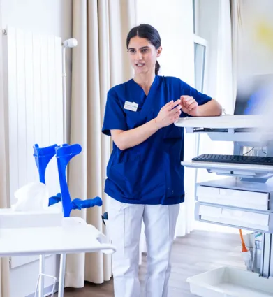 Pflegekraft in blauer Uniform steht in einem Klinikraum neben einer Sterilgut- und Messanlage, ordnet Instrumente an und richtet sich an einem Tisch mit medizinischen Geräten.