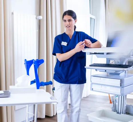 Pflegekraft in blauer Uniform steht in einem Klinikraum neben einer Sterilgut- und Messanlage, ordnet Instrumente an und richtet sich an einem Tisch mit medizinischen Geräten.