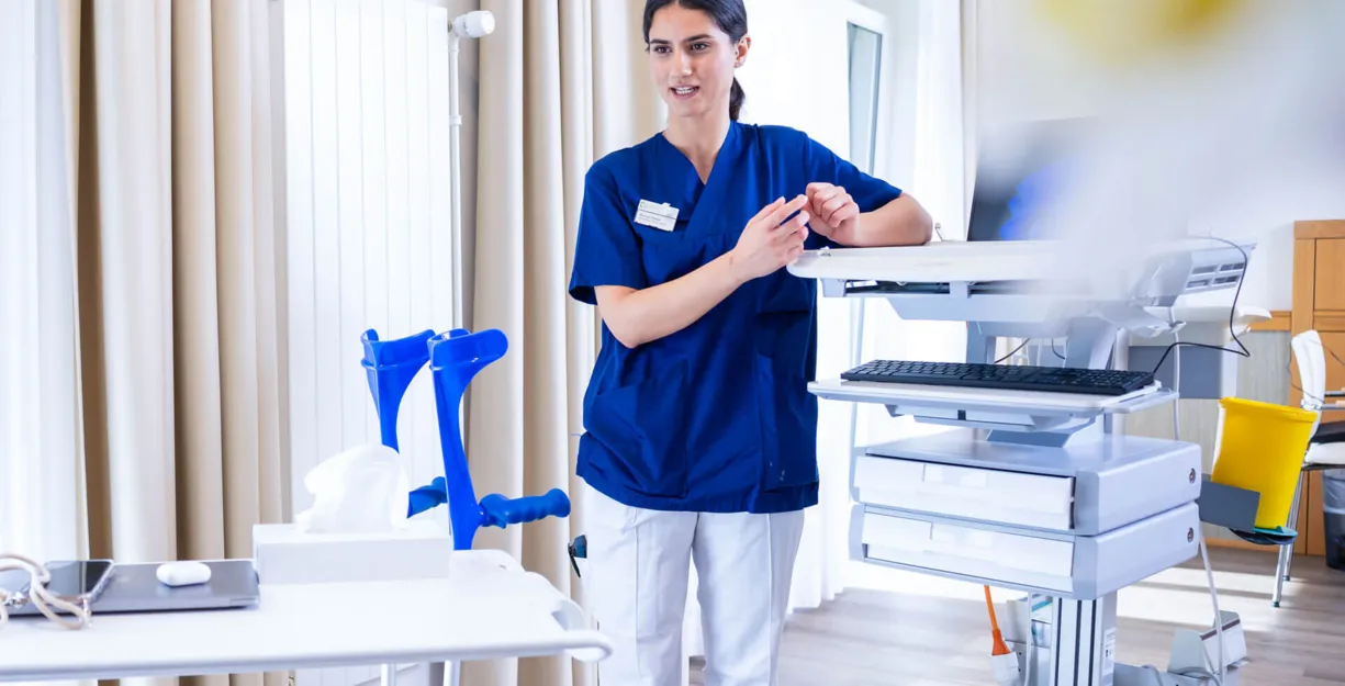 Pflegekraft in blauer Uniform steht in einem Klinikraum neben einer Sterilgut- und Messanlage, ordnet Instrumente an und richtet sich an einem Tisch mit medizinischen Geräten.