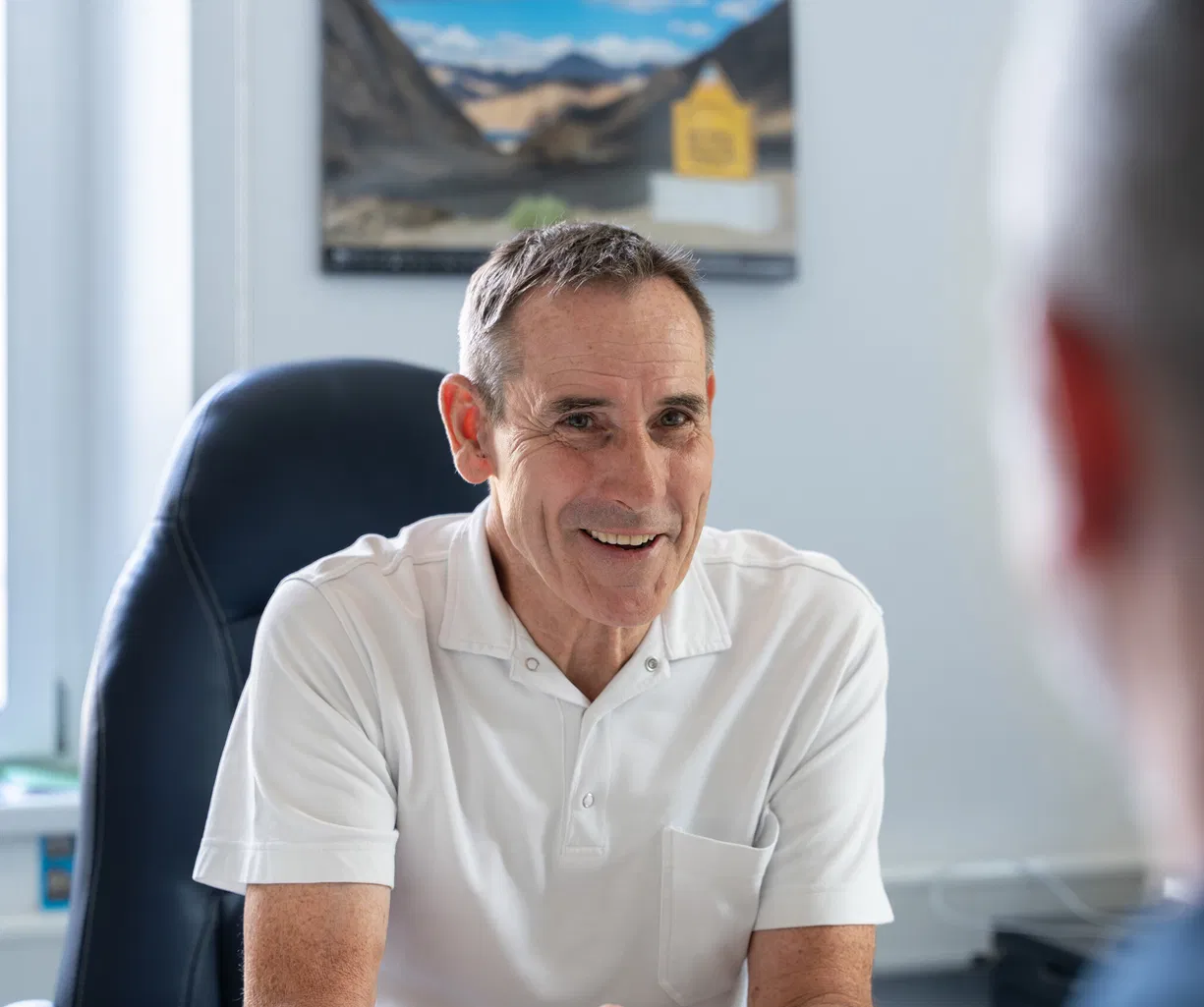Lächelnder Mann im weißen Poloshirt sitzt einem Gesprächspartner in einem Büro gegenüber. Im Hintergrund sind ein Fenster, ein Sessel und ein gerahmtes Landschaftsbild zu sehen.