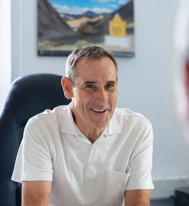 Lächelnder Mann im weißen Poloshirt sitzt einem Gesprächspartner in einem Büro gegenüber. Im Hintergrund sind ein Fenster, ein Sessel und ein gerahmtes Landschaftsbild zu sehen.