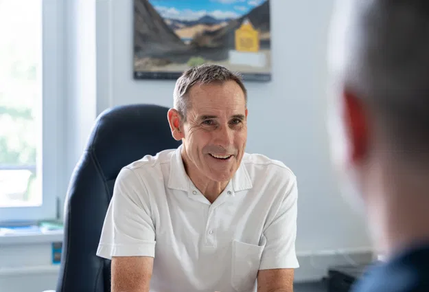 Lächelnder Mann im weißen Poloshirt sitzt einem Gesprächspartner in einem Büro gegenüber. Im Hintergrund sind ein Fenster, ein Sessel und ein gerahmtes Landschaftsbild zu sehen.