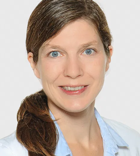 Porträt einer lächelnden Frau mit braunem Pferdeschwanz, hellblauem Hemd und weißem Kittel vor hellem Hintergrund.