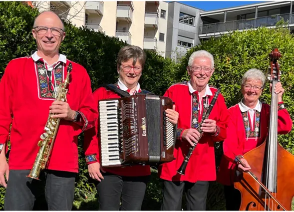 Vier Musiker in roten Trachtenhemden stehen im Freien vor einer grünen Hecke. Sie halten verschiedene Instrumente: ein Saxophon, ein Akkordeon, eine Klarinette und ein Kontrabass. Im Hintergrund sind moderne Wohngebäude und ein blauer Himmel sichtbar.