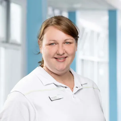 Frau mit braunen Haaren und lächelndem Gesicht, trägt ein weißes Polohemd mit einem Namensschild, im Hintergrund helle, moderne Innenarchitektur mit blauen Akzenten.