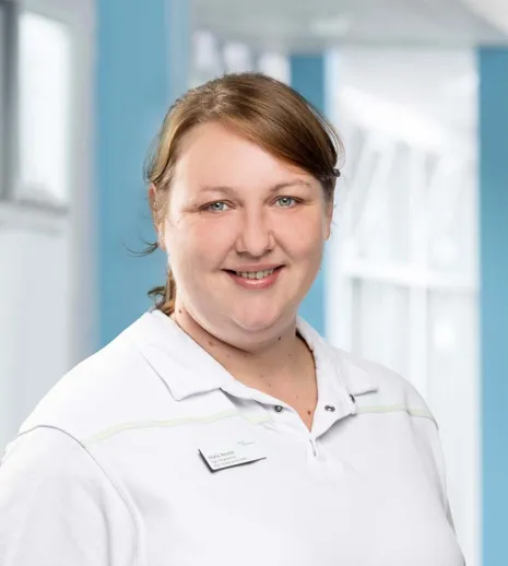 Frau mit braunen Haaren und lächelndem Gesicht, trägt ein weißes Polohemd mit einem Namensschild, im Hintergrund helle, moderne Innenarchitektur mit blauen Akzenten.