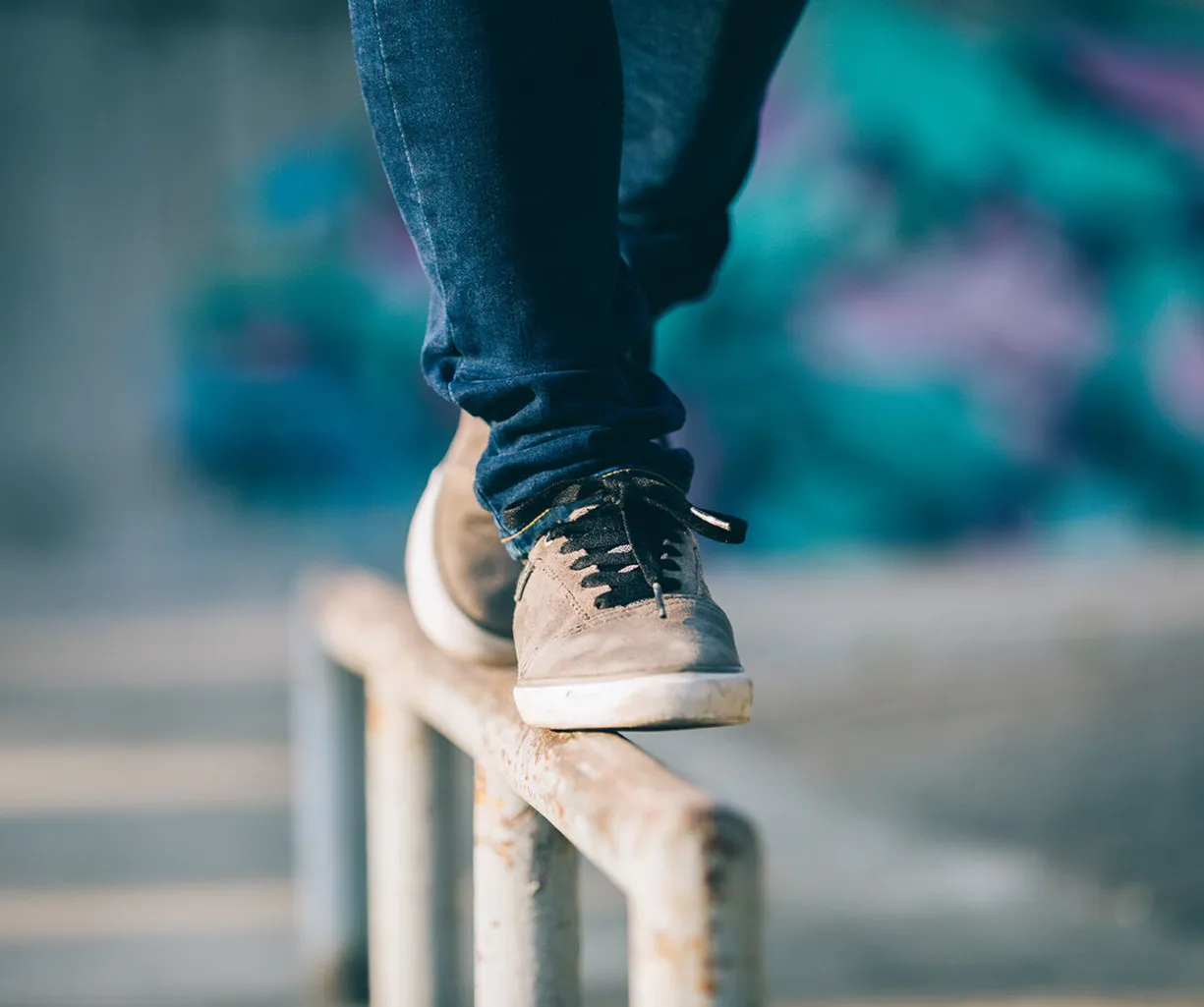 Ein Fuß in einem grauen Sneaker balanciert auf einem Metallgeländer, während im Hintergrund eine farbige Wand mit Graffiti sichtbar ist.