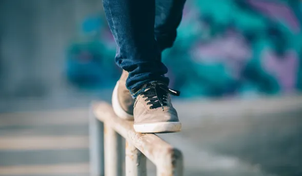 Ein Fuß in einem grauen Sneaker balanciert auf einem Metallgeländer, während im Hintergrund eine farbige Wand mit Graffiti sichtbar ist.