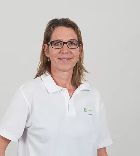Porträt einer lächelnden Frau mit Brille und schulterlangem Haar in weißem Polohemd vor hellgrauem Hintergrund.