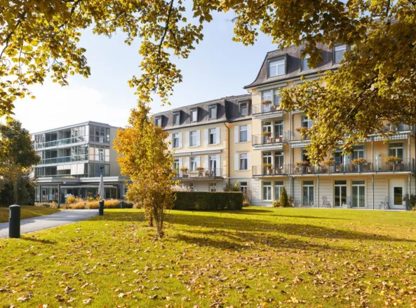 Zwei mehrstöckige Gebäude in einem Park mit herbstlichen Bäumen und buntem Laub. Im Vordergrund eine grüne Wiese mit verstreuten Blättern und einem Weg, der zu den Gebäuden führt.